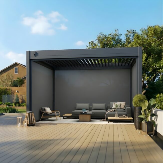 A 3x4m dark grey louvered pergola stands on a light-colored wooden deck. The pergola features three pulled-down privacy blinds and a slatted roof. Underneath, a modern grey sectional sofa and a low wooden coffee table create an outdoor living area. The background shows a clear blue sky, green trees, and the corner of a residential house