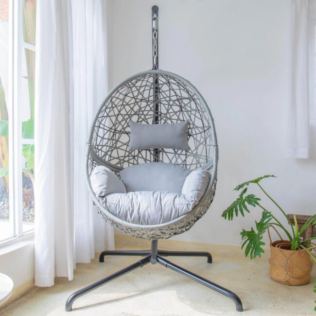 Modern rattan hanging chair with soft cushions, placed in a bright indoor space with natural light filtering through white curtains and houseplants nearby.